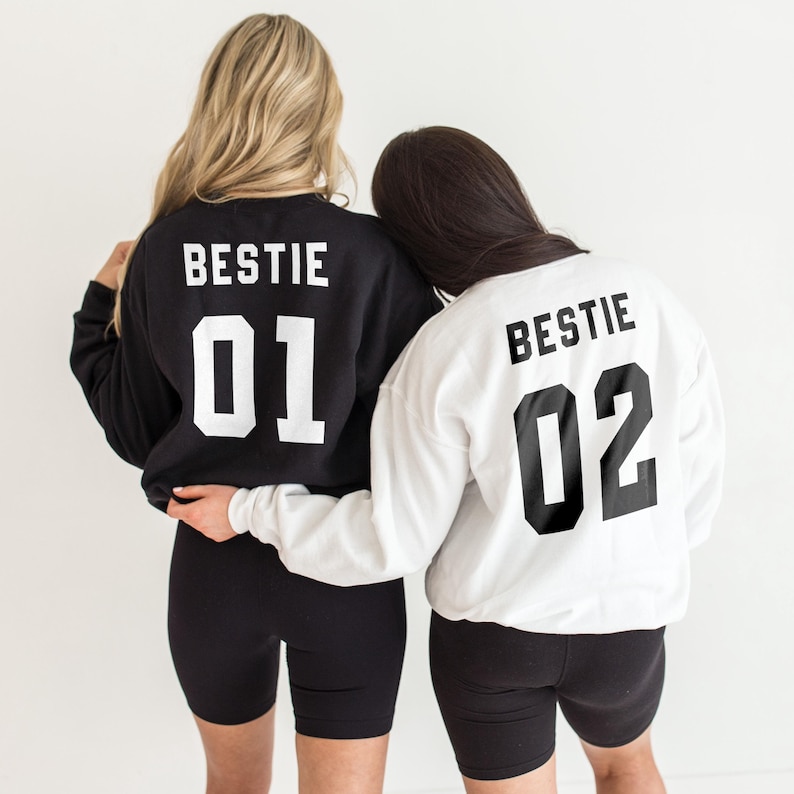 Puede incluir: Dos sudaderas blancas y negras con la palabra "Bestie" y los n&uacute;meros "01" y "02" impresos en la espalda. Las sudaderas las llevan dos personas de pie espalda con espalda.