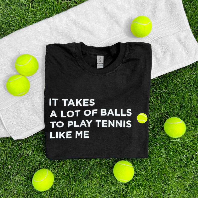 Op de afbeelding: Zwart T-shirt met witte tekst die luidt "Het kost veel ballen om tennis te spelen zoals ik". Een gele tennisbal bevindt zich naast de tekst. Het T-shirt is omringd door gele tennisballen op een groene grasmat.