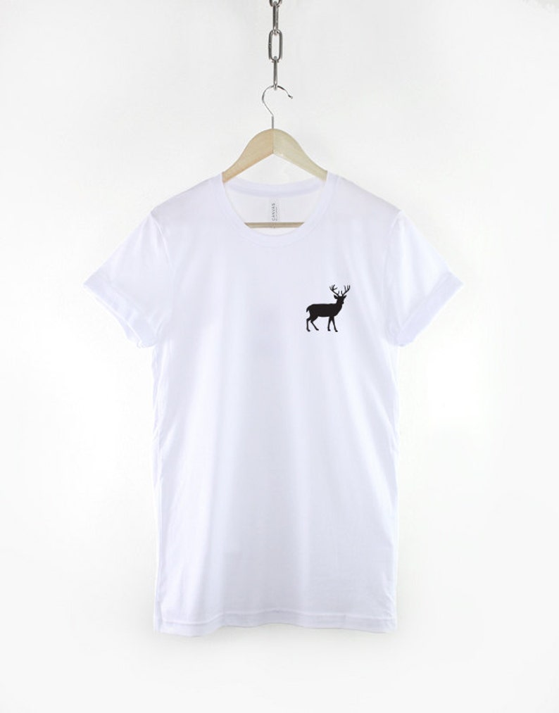 Puede incluir: Una camiseta blanca de manga corta con un peque&ntilde;o gr&aacute;fico de silueta negra de un ciervo con astas situado en la parte superior izquierda del pecho. La prenda se muestra en una percha de madera colgada de una cadena met&aacute;lica sobre fondo liso.