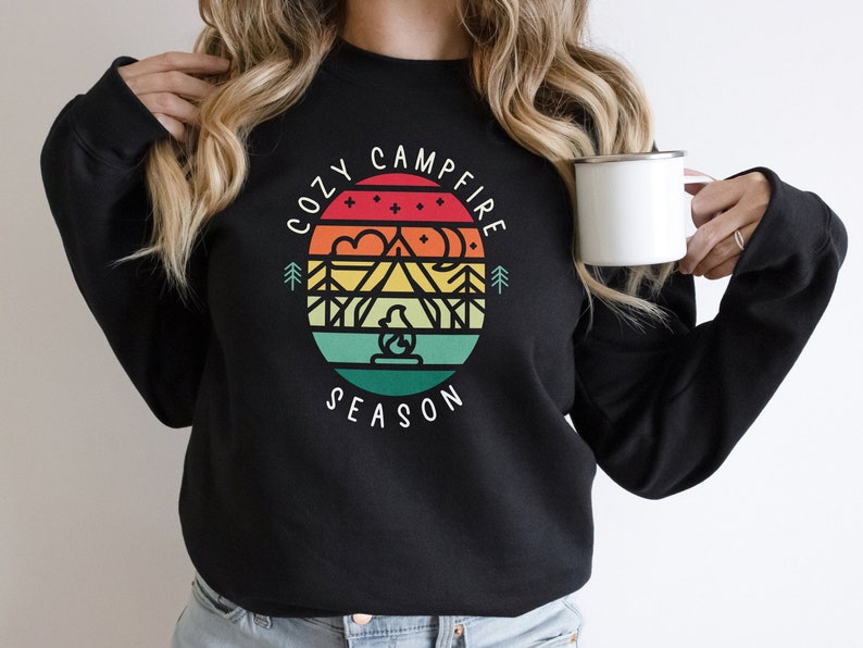 Op de afbeelding: Zwart sweatshirt met een retro-grafisch ontwerp met een berglandschap, een kampvuur en de tekst "Cozy Campfire Season".