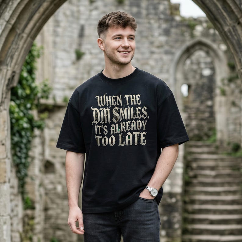 May include: A black short-sleeved t-shirt featuring the text "WHEN THE DM SMILES, IT'S ALREADY TOO LATE" printed in a distressed, gothic-style font across the chest. The shirt has a crew neckline and a relaxed fit, worn with dark gray jeans.