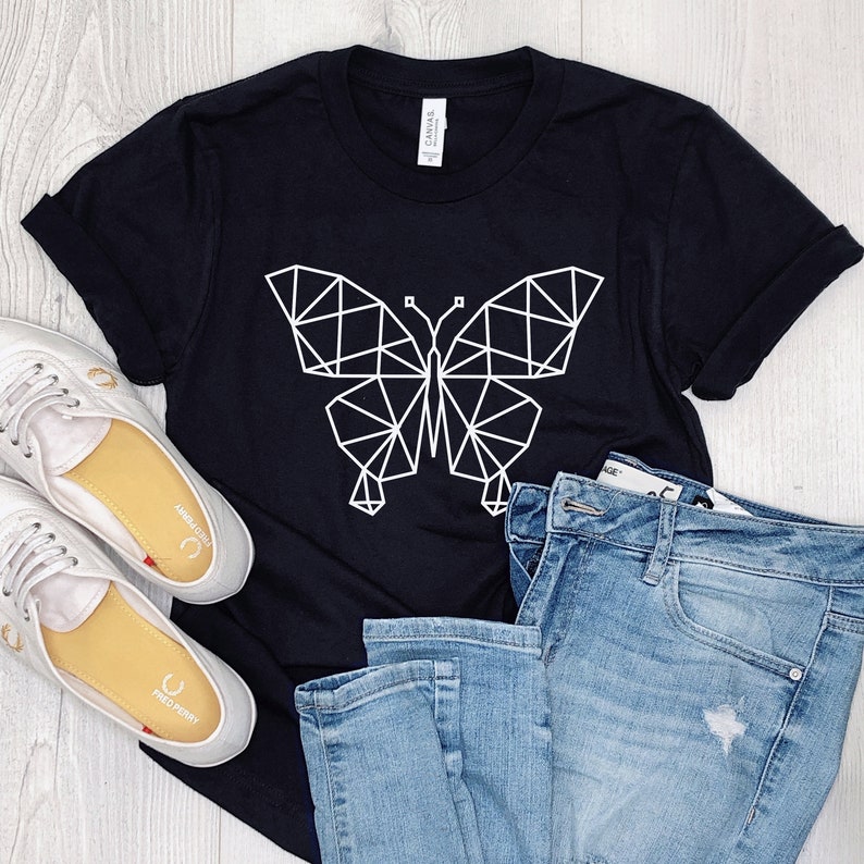 May include: A black t-shirt with a white geometric butterfly design. The shirt is laying on a white wooden surface with a pair of white sneakers and blue jeans.