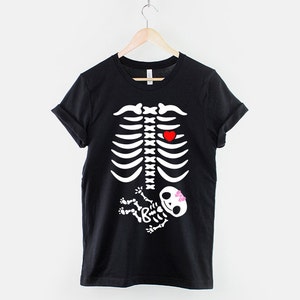 May include: Black t-shirt with a white skeleton graphic. The skeleton has a red heart in its chest and a small white skeleton baby in its belly with a pink bow.