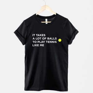 Op de afbeelding: Zwart T-shirt met witte tekst die luidt "Het kost veel lef om tennis te spelen zoals ik" en een geel tennisbal-icoon.