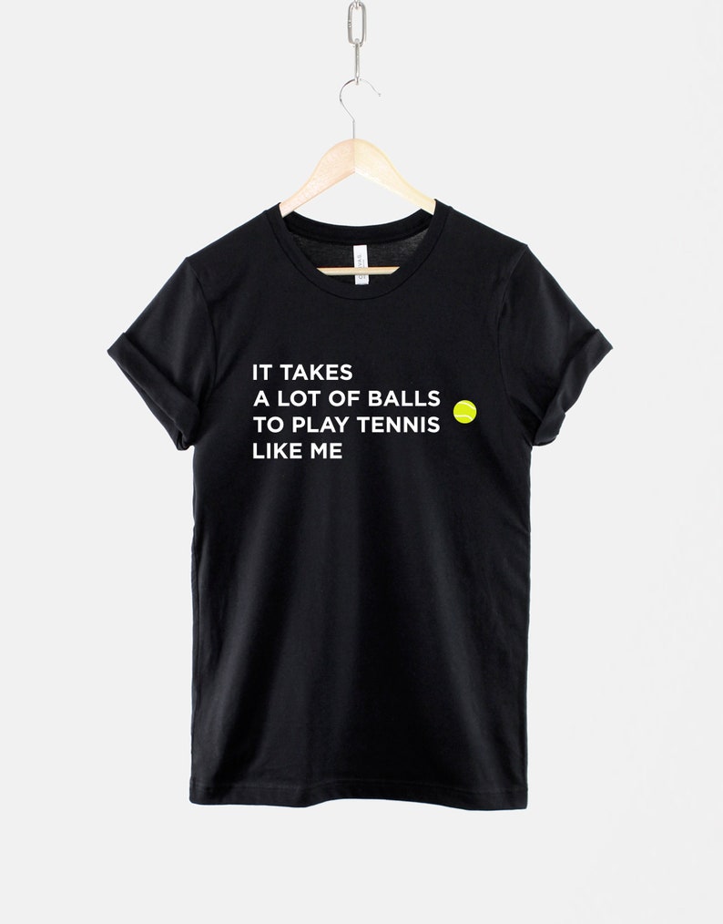 Op de afbeelding: Zwart T-shirt met witte tekst die luidt "Het kost veel lef om tennis te spelen zoals ik" en een geel tennisbal-icoon.