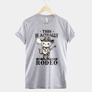 Peut inclure: T-shirt gris avec un chat de dessin anim&eacute; portant un chapeau de cowboy et jouant de la guitare. Le texte sur le t-shirt est "This is actually my first rodeo."