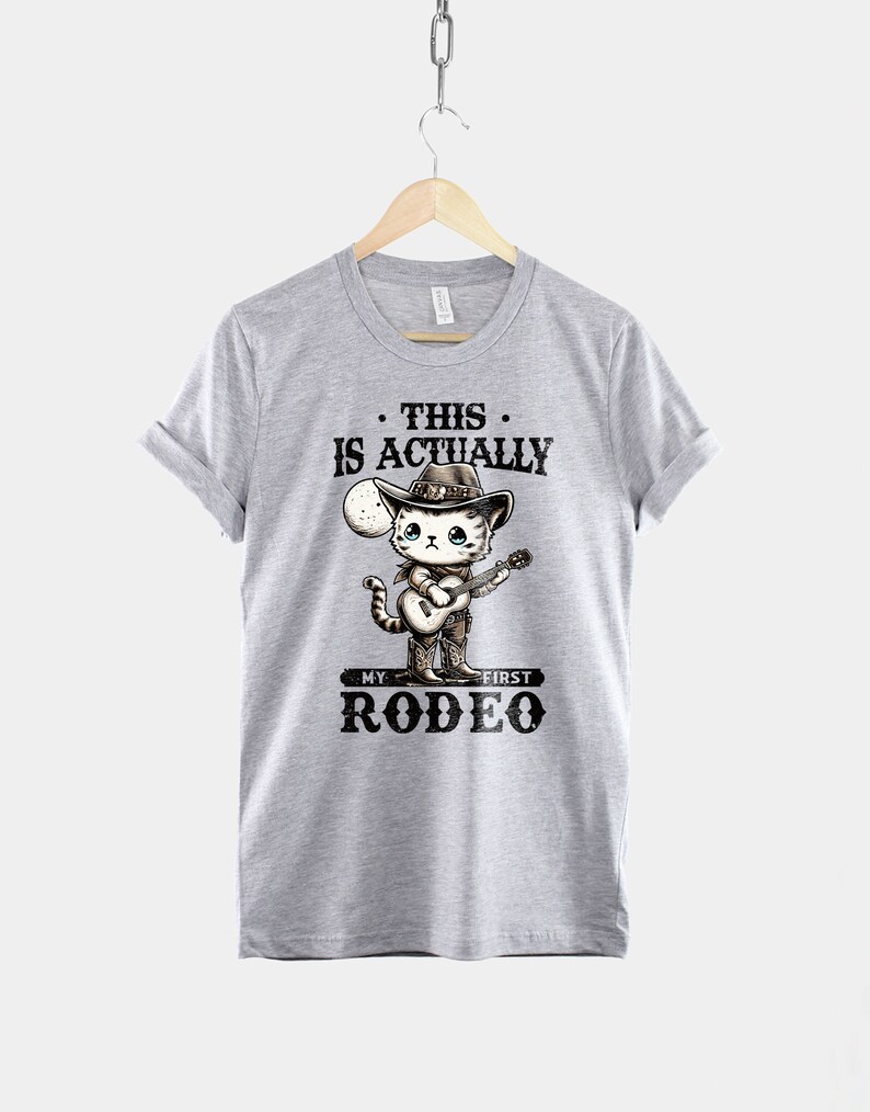 Peut inclure: T-shirt gris avec un chat de dessin anim&eacute; portant un chapeau de cowboy et jouant de la guitare. Le texte sur le t-shirt est "This is actually my first rodeo."
