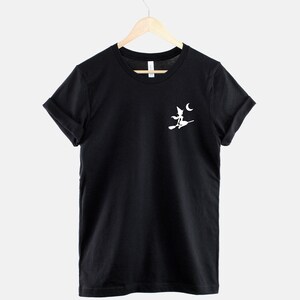 Op de afbeelding: Zwart T-shirt met een wit silhouet van een heks die op een bezem vliegt met een halve maan op de achtergrond.