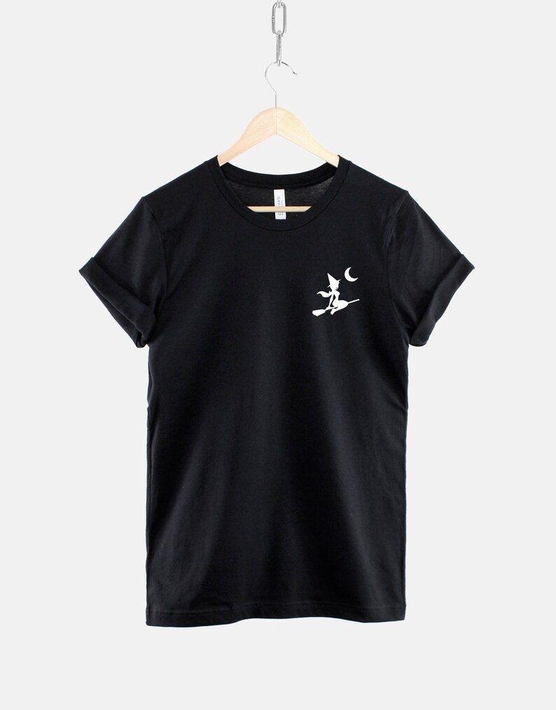 Op de afbeelding: Zwart T-shirt met een wit silhouet van een heks die op een bezem vliegt met een halve maan op de achtergrond.