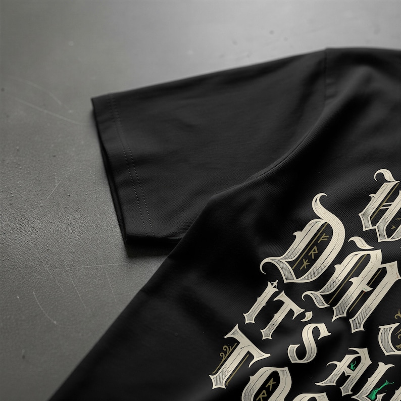 May include: A black short-sleeved t-shirt featuring decorative, light-colored gothic-style lettering printed on the front. The text includes the words "IT'S ALL" in a stylized, ornate font with intricate details and small green accents.