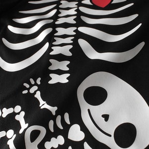 May include: Black t-shirt with a white skeleton design. The skeleton has a red heart in its chest and a baby skeleton inside its ribcage.
