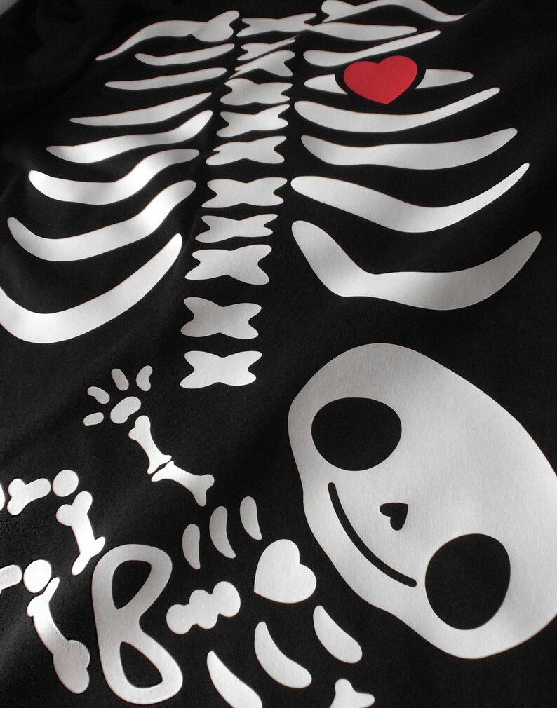 May include: Black t-shirt with a white skeleton design. The skeleton has a red heart in its chest and a baby skeleton inside its ribcage.