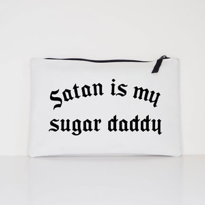 Op de afbeelding: Witte canvas toilettas met zwarte tekst die "Satan is my sugar daddy" zegt.