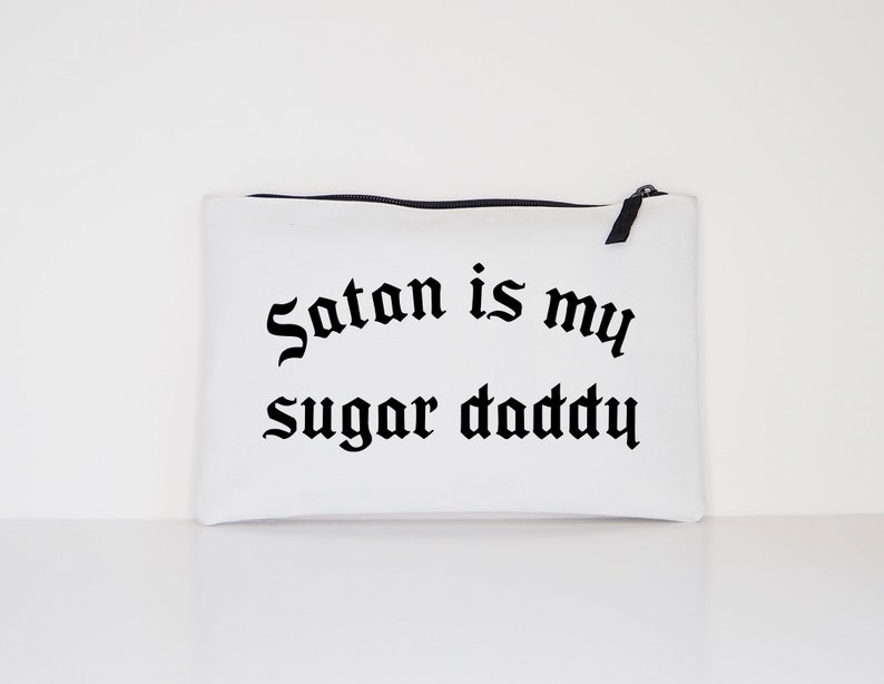 Op de afbeelding: Witte canvas toilettas met zwarte tekst die "Satan is my sugar daddy" zegt.