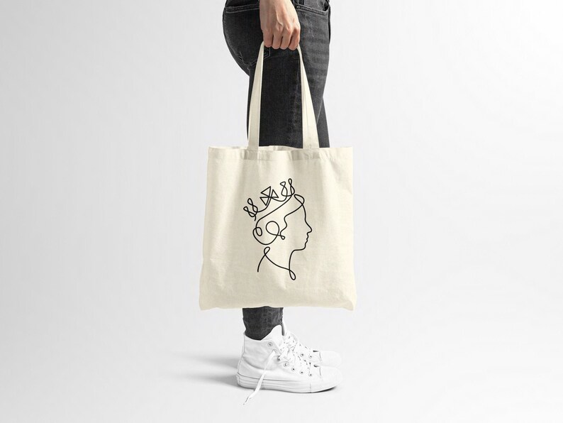 Op de afbeelding: Een witte canvas tote bag met een zwarte lijntekening van een vrouwenhoofd met een kroon op haar hoofd.
