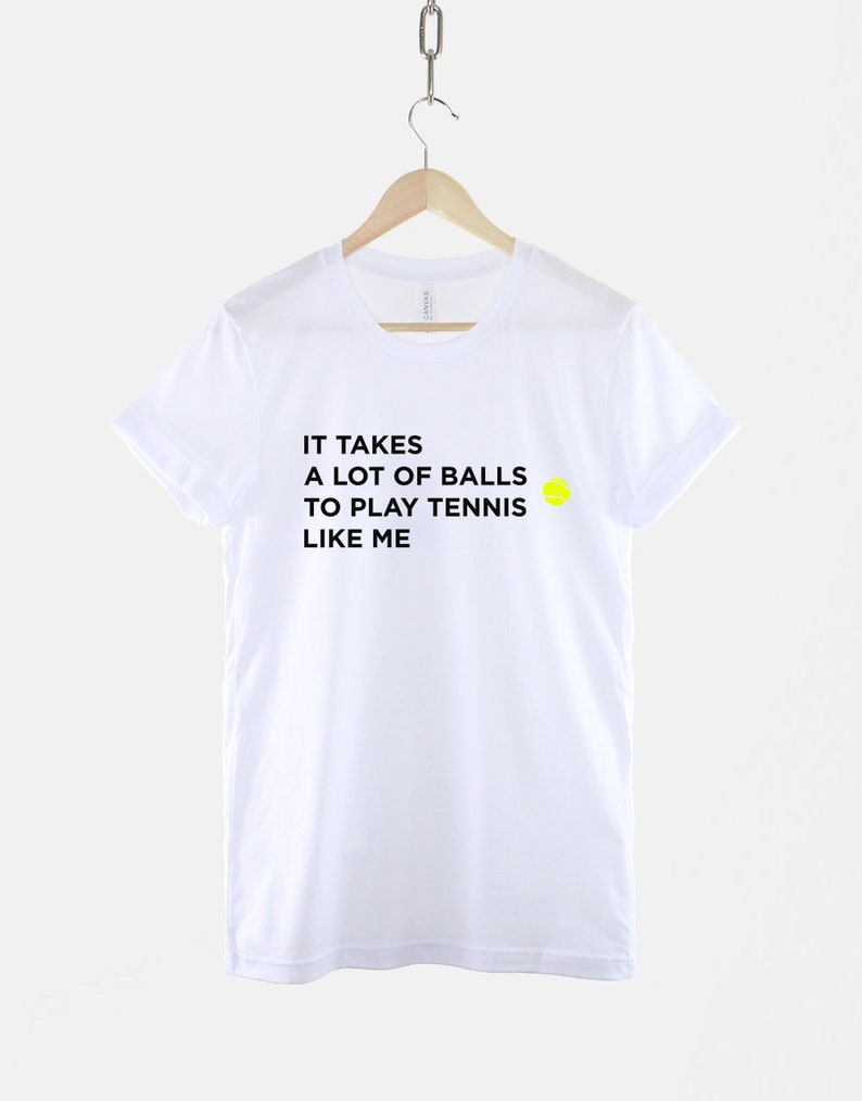 Op de afbeelding: Wit T-shirt met zwarte tekst die luidt "Het kost veel ballen om tennis te spelen zoals ik" en een gele tennisbal afbeelding.