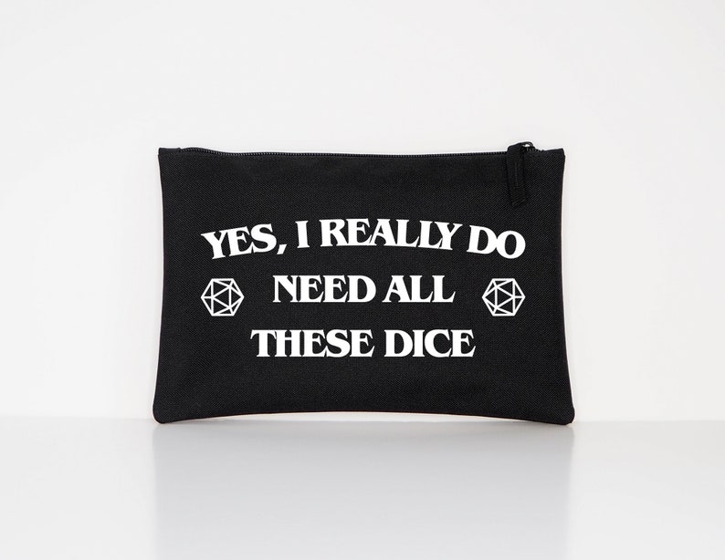 Puede incluir: Bolsa rectangular negra con cierre de cremallera. Texto blanco en la parte delantera que dice "YES, I REALLY DO NEED ALL THESE DICE", con dos formas geom&eacute;tricas blancas. La bolsa es de tela.
