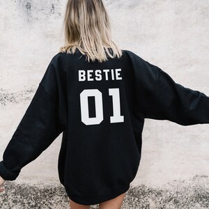 Puede incluir: Sudadera negra con las palabras "BESTIE 01" impresas en la espalda en blanco.