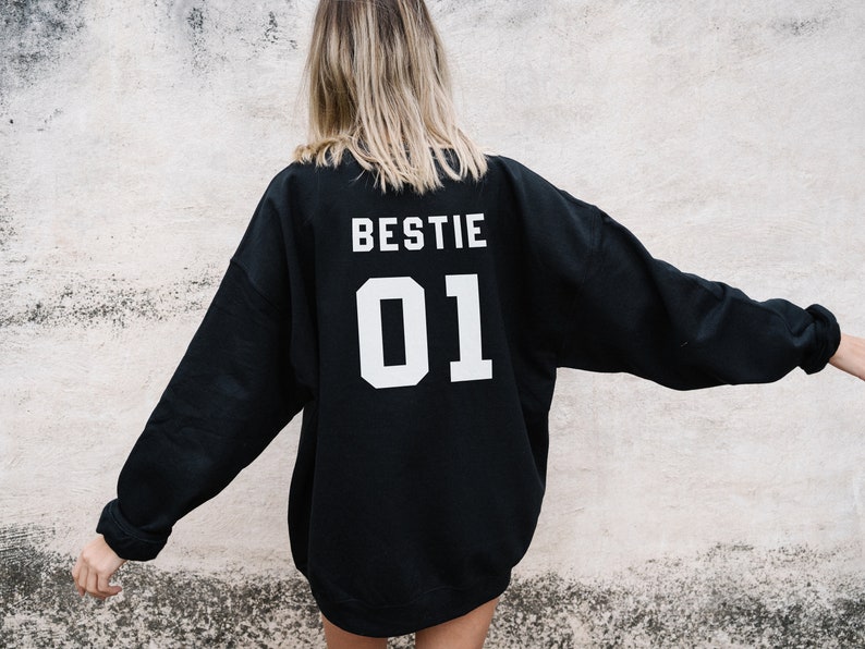 Puede incluir: Sudadera negra con las palabras "BESTIE 01" impresas en la espalda en blanco.