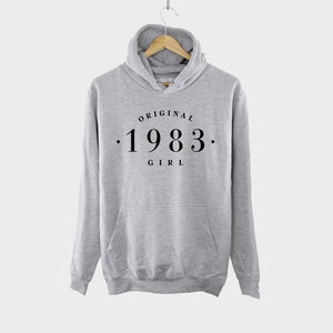 K&ouml;nnte beinhalten: Grauer Kapuzenpullover mit dem Aufdruck "ORIGINAL . 1983 . GIRL" auf der Vorderseite.