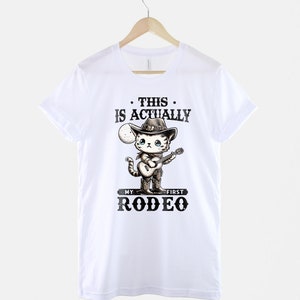 Peut inclure: T-shirt blanc avec un chat de dessin anim&eacute; portant un chapeau de cowboy et jouant de la guitare. Le texte sur le t-shirt est "This is actually my first rodeo."