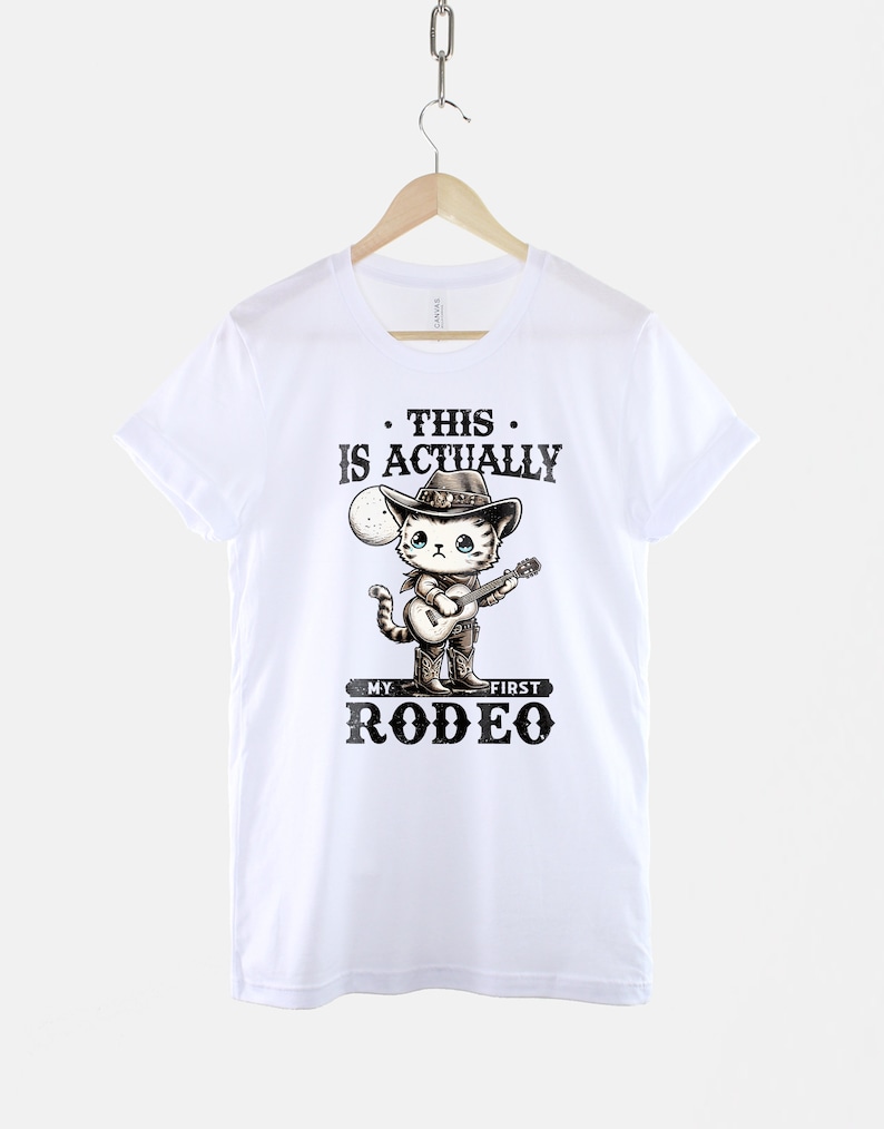 Peut inclure: T-shirt blanc avec un chat de dessin anim&eacute; portant un chapeau de cowboy et jouant de la guitare. Le texte sur le t-shirt est "This is actually my first rodeo."