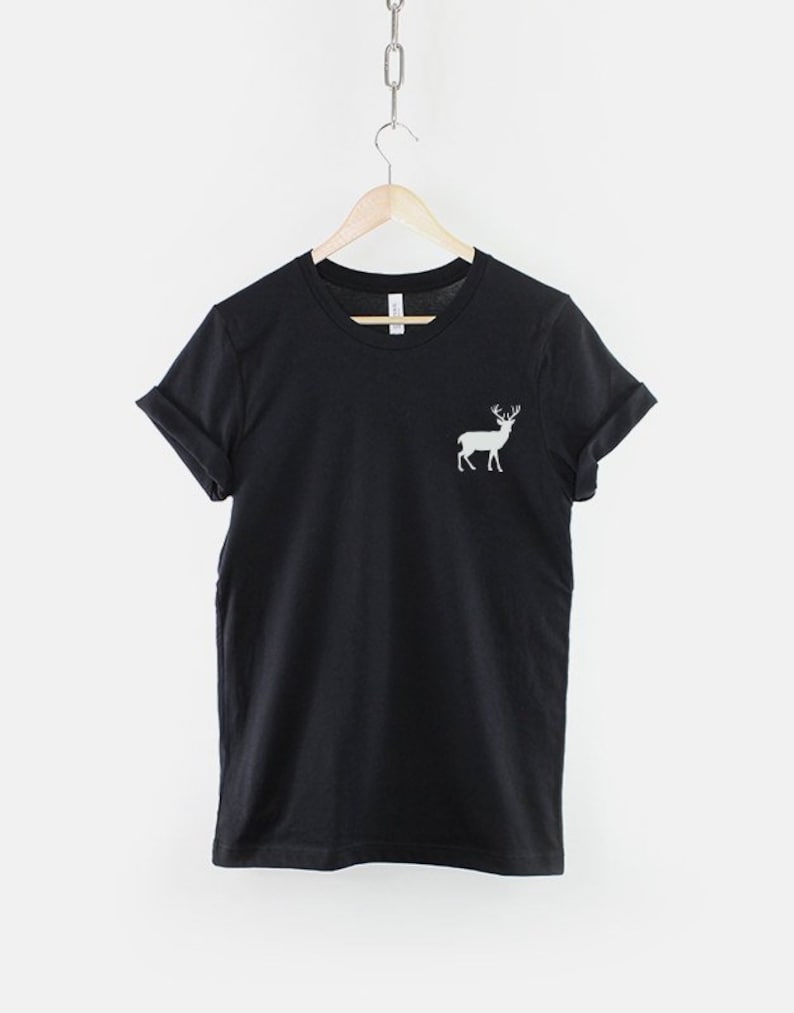 Puede incluir: Una camiseta negra de manga corta con cuello redondo y mangas remangadas. La prenda presenta un peque&ntilde;o gr&aacute;fico blanco de un ciervo de pie con astas situado en la parte superior izquierda del pecho. Se muestra en una percha de madera.