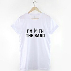 Puede incluir: Camiseta blanca con texto negro que dice "I'm with the band" y un gesto de mano rock and roll negro.