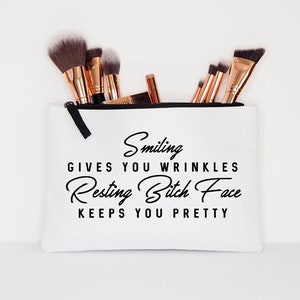 May include: White makeup bag with black zipper and text that reads "Smiling gives you wrinkles, Resting Bitch Face keeps you pretty." The bag contains several makeup brushes with black bristles and rose gold handles.