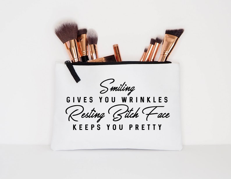 May include: White makeup bag with black zipper and text that reads "Smiling gives you wrinkles, Resting Bitch Face keeps you pretty." The bag contains several makeup brushes with black bristles and rose gold handles.