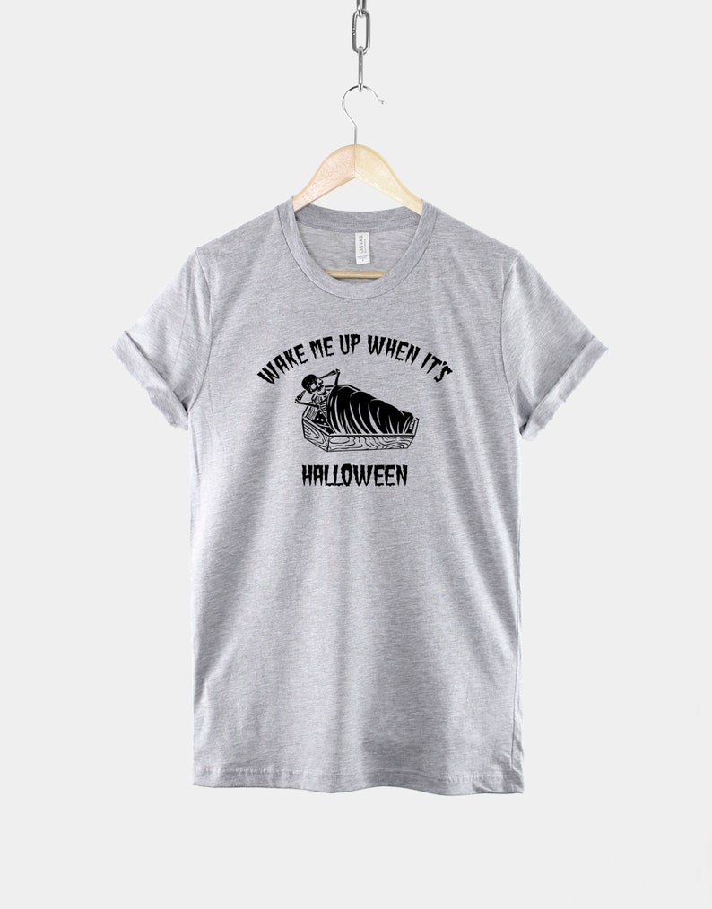 Puede incluir: Camiseta gris con un gr&aacute;fico negro de un esqueleto en un ata&uacute;d. El texto dice "Despi&eacute;rtame cuando sea Halloween".