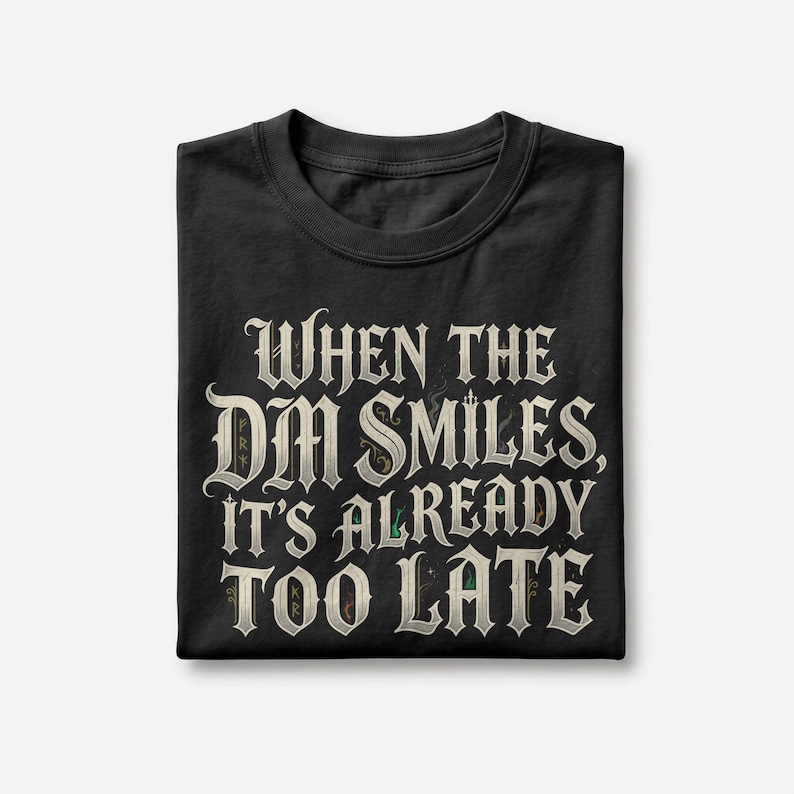 May include: A black short-sleeved t-shirt featuring the white text "WHEN THE DM SMILES, IT'S ALREADY TOO LATE" in a decorative, gothic-inspired font. The lettering has subtle, intricate details within the characters and a clean, folded layout.