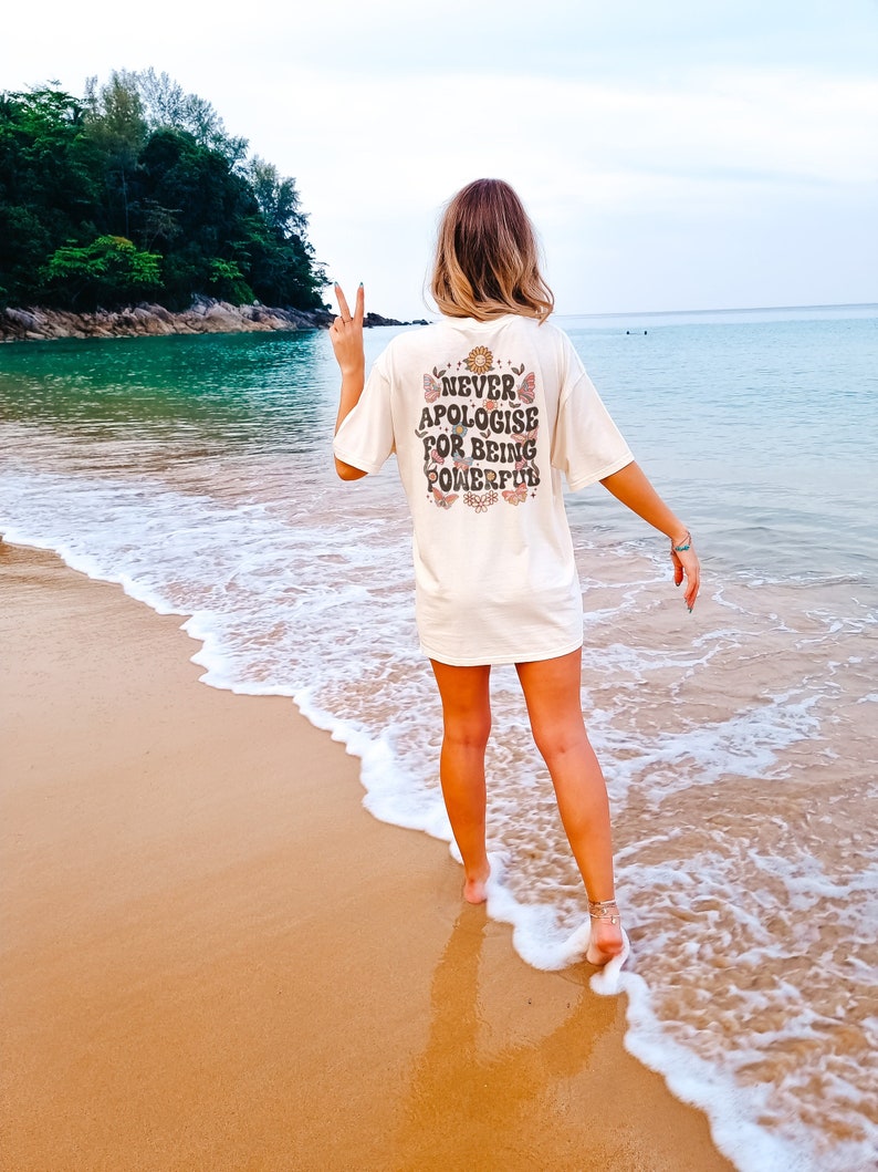 May include: A woman wearing a white t-shirt with the text "NEVER APOLOGISE FOR BEING POWERFUL" printed on the back. She is standing on a sandy beach with the sea in the background.