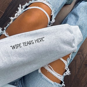 May include: A gray sweatshirt with the text "Wipe Tears Here" printed on it. The sweatshirt is being worn by a person wearing ripped denim jeans.