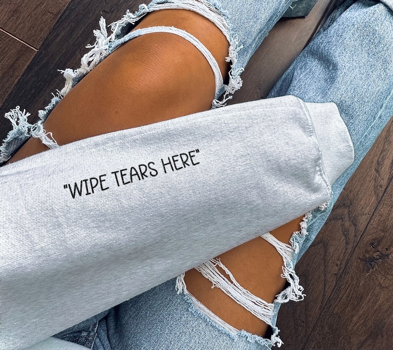 May include: A gray sweatshirt with the text "Wipe Tears Here" printed on it. The sweatshirt is being worn by a person wearing ripped denim jeans.