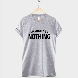 May include: A heather gray t-shirt with the phrase "THANKS FOR NOTHING" printed in bold black letters. The short-sleeved shirt is hanging on a wooden hanger.