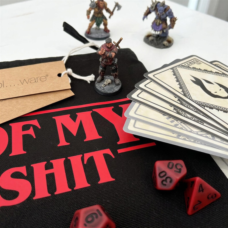 May include: A black fabric drawstring bag with the red text OF MY SHIT, accompanied by three painted miniature fantasy figures, a stack of rectangular game cards with a flame icon, and two red multi-sided dice with black numbers.