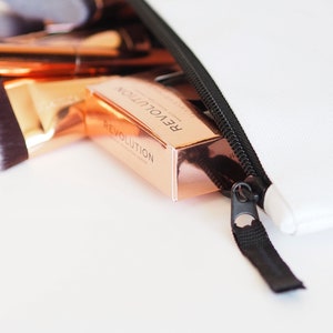 May include: A white zippered pouch with a black zipper pull. The pouch is partially open, revealing a rose gold makeup brush set and a rose gold box with the word "REVOLUTION" printed on it.
