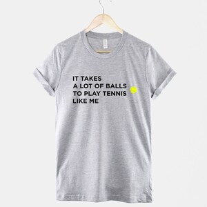 Op de afbeelding: Een grijs T-shirt met zwarte tekst die luidt "Het kost veel lef om tennis te spelen zoals ik" en een geel tennisbal-icoon.