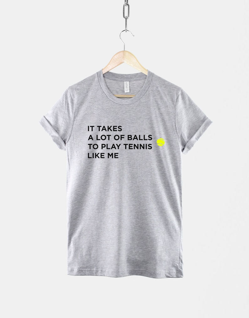 Op de afbeelding: Een grijs T-shirt met zwarte tekst die luidt "Het kost veel lef om tennis te spelen zoals ik" en een geel tennisbal-icoon.