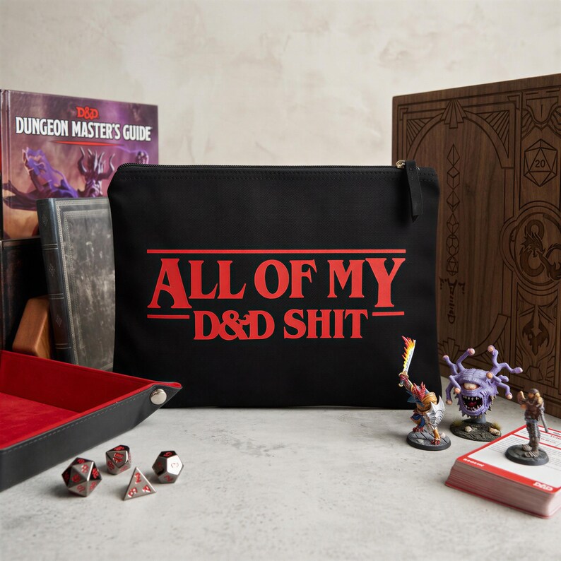 May include: A black rectangular zippered pouch featuring the red text ALL OF MY D&D SHIT printed on the front. The pouch is positioned among various tabletop gaming accessories, including a red-lined tray, polyhedral dice, and miniature figures.
