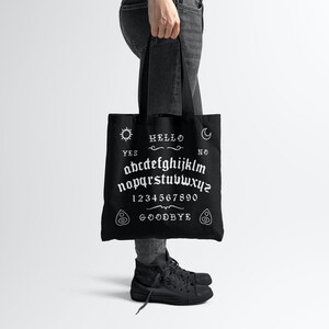 May include: Black tote bag with a white ouija board design. The design includes the letters of the alphabet, numbers 1-9 and 0, the words "yes", "no", "hello", and "goodbye", and symbols for a sun, moon, and a hand.