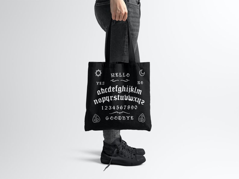 May include: Black tote bag with a white ouija board design. The design includes the letters of the alphabet, numbers 1-9 and 0, the words "yes", "no", "hello", and "goodbye", and symbols for a sun, moon, and a hand.