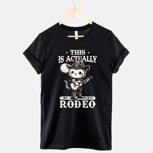 Peut inclure: T-shirt noir avec un graphique blanc d'un chat portant un chapeau de cowboy et jouant de la guitare. Le texte sur le t-shirt est "This is actually my first rodeo."