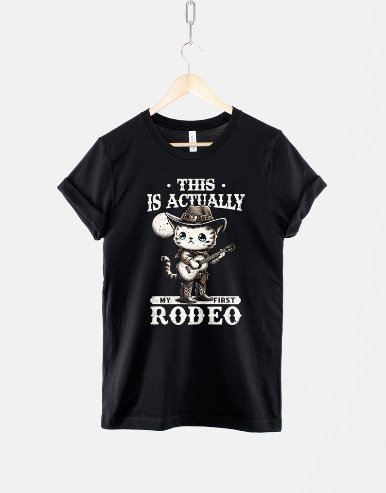 Peut inclure: T-shirt noir avec un graphique blanc d'un chat portant un chapeau de cowboy et jouant de la guitare. Le texte sur le t-shirt est "This is actually my first rodeo."