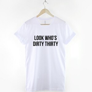 Może przedstawiać: Biała koszulka z czarnym napisem "Look Who's Dirty Thirty".