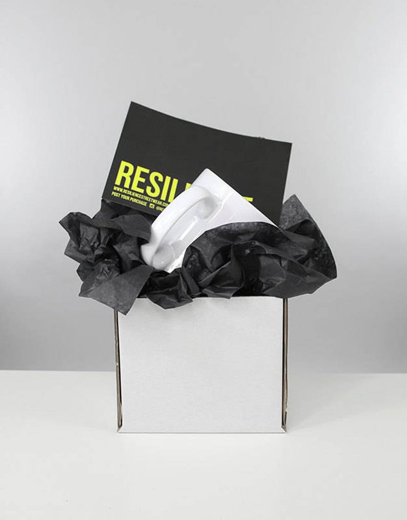 May include: A white ceramic mug with a black and yellow logo that says "RESILIENT" and the website address "www.resilientstreetwear.com". The mug is packaged in a white box with black tissue paper.