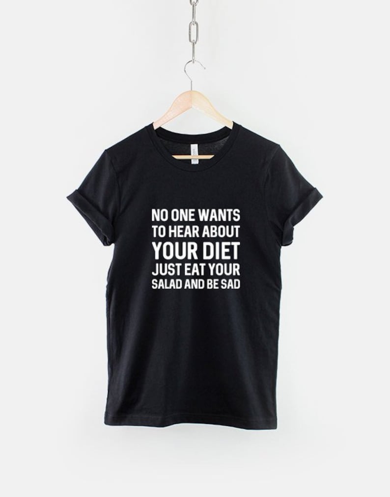 May include: Black t-shirt with white text that reads "NO ONE WANTS TO HEAR ABOUT YOUR DIET JUST EAT YOUR SALAD AND BE SAD"