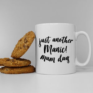 May include: White ceramic mug with black text that reads "just another Manic! mum day".  The mug is sitting on a white surface next to three chocolate chip cookies.