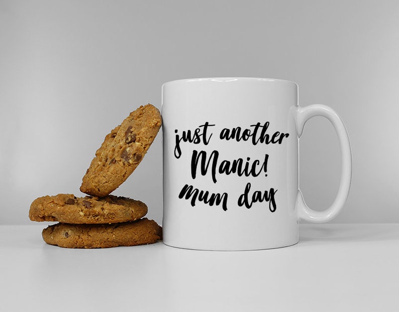 May include: White ceramic mug with black text that reads "just another Manic! mum day".  The mug is sitting on a white surface next to three chocolate chip cookies.